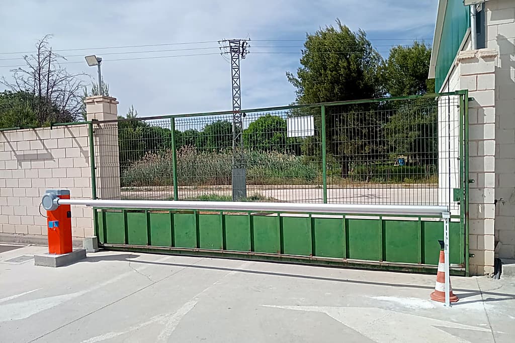Barrera automática instalada junto a una puerta corredera metálica verde para controlar el acceso de vehículos.